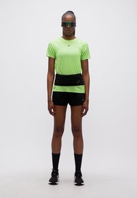 Γυναίκα που φοράει νεον πράσινο αθλητικό πουκάμισο, μαύρο σορτς, μαύρη ζώνη τρεξίματος, μαύρες κάλτσες και αντανακλαστικά αθλητικά γυαλιά ηλίου, στέκεται μπροστά σε λευκό φόντο.