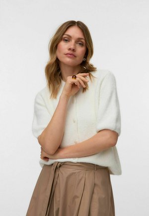 Žena so svetlými blond vlasmi po ramená nosí biely krátkym rukávom pletený top, béžovú sukňu s vysokým pásom, jantárový prsteň a strieborné kruhové náušnice.