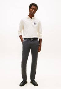 Polo blanc à manches longues avec logo brodé, associé à un pantalon gris slim et des chaussures noires. Fond uni.