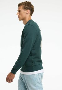 Felpa verde a coste con colletto rotondo, polsini aderenti e un logo discreto sul retro, abbinata a jeans in denim blu chiaro.
