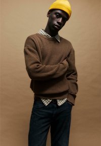 Brązowy sweter w prążki nałożony na koszulę w kratę, ciemne dżinsy i żółty beanie. Gładkie tekstury i nałożone wzory tworzą głębię.