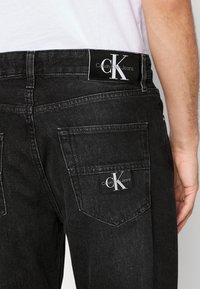 Svarta denimjeans med en Calvin Klein midjebandsetikett, två bakfickor och en strukturerad yta. Minimalistisk design med diskret varumärkesprofil.