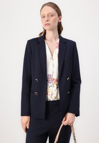 Námořnický modrý dvouřadý blazer se zlatými akcenty a odpovídajícími kalhotami, vrstvený přes bílou blůzu s květinovým potiskem. Hladká textura látky.