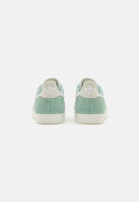 Zapatillas de ante verde con una textura suave y suela de goma blanca. Presentan una pestaña blanca en el talón y un logo de la marca en la parte trasera. Diseño sencillo.