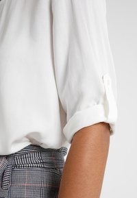 Vit blus med uppvikta ärmar och knappdetalj samt rutiga byxor i midjan på en arm med medelbrun hudton.