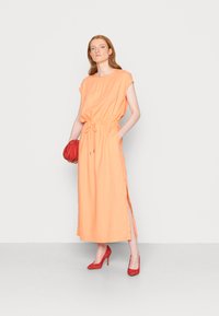 Vestido longo, laranja, sem mangas, com um cinto ajustável na cintura e fendas laterais, combinado com sapatos de salto alto vermelhos e uma pequena bolsa vermelha.