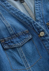 Veste en denim bleu vue de face avec rabat de poche poitrine visible, coutures surpiquées et un bouton en métal portant l'inscription « Next Denim Since 1982 ».