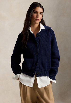 Kvinde iført en mørkeblå knappet cardigan over en hvid skjorte med krave og beige bukser, poserer foran en neutral baggrund.