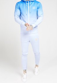 Uomo in felpa con zip blu sfumata e pantaloni della tuta coordinati, con scarpe da ginnastica bianche, in piedi davanti a uno sfondo bianco uniforme.