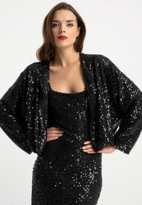 Czarna cekinowa kurtka o luźnym kroju i szerokich rękawach, zestawiona z dopasowaną cekinową sukienką o tej samej fakturze, obie z połyskującą teksturą.