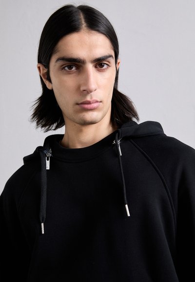 Črna kapuca z teksturirano površino, ki ima vrvico z metalnimi zaključki in raglan rokave. Kapuca ima visok ovratnik.