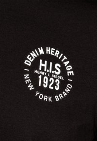 Černá látka se zaobleným bílým logem, na němž je nápis "DENIM HERITAGE H.I.S HENRY I. SIEGEL 1923 NEW YORK BRAND."