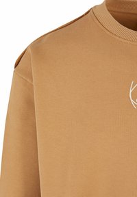 Brun bomulds sweatshirt med ribbet krave og skulderdetaljer, der har et broderet hvidt logo på fronten.