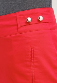 Shorts en coton rouge avec une taille plate, fermeture à deux boutons argentés et des coutures nettes, avec un détail de poche latérale.