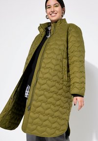 Cappotto lungo imbottito verde oliva con motivo testurizzato, chiusura a pressione e tasche laterali. Indossato sopra un maglione nero e una camicia a quadri.