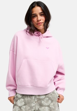 Jeune femme aux cheveux foncés portant un sweat à capuche rose clair et un pantalon gris à motifs floraux, debout devant un fond blanc uni.