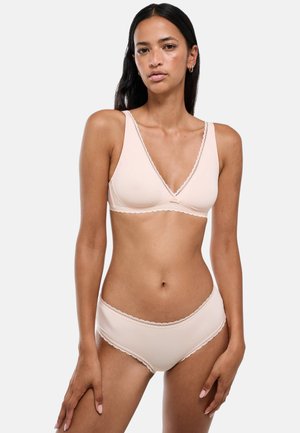 Femme portant un soutien-gorge rose clair orné de dentelle et une culotte taille haute assortie, se tenant devant un fond clair uni.