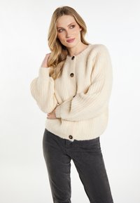 Cardigan in maglia a treccia color crema con scollo rotondo, tre grandi bottoni scuri e maniche a sbuffo, abbinato a jeans neri. Texture morbida.