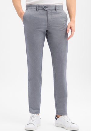 Man die slim-fit grijze broek en witte sneakers draagt, met één hand in zijn zak staand tegen een effen achtergrond.