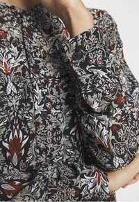 Mustalilakkinen kukkapusero, jossa on monimutkaisia kuvioita punaisena, ivorynä ja harmaana. Kappaleessa on kaareva kaulus, nappisuljenta edessä ja teksturoitu kangas.