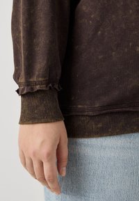 Felpa marrone scuro con macchie più chiare, polsini a costina e scollatura arricciata. Il tessuto sembra morbido con una consistenza leggermente usurata.