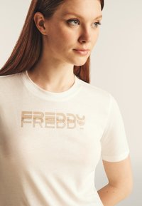 Camiseta blanca de algodón con un gran logo de Freddy en metal dorado y la impresión "MOOD", de manga corta y cuello redondo. Textura suave.
