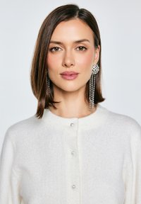 Bílé pletené kardigany s kulatým výstřihem, který zdobí perlové knoflíky. Dlouhé třpytivé náušnice s kaskádovým designem dotvářejí vzhled.