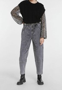 Maglione nero lavorato a maglia con un motivo a treccia sopra una blusa trasparente a maniche lunghe con stampa animalier. Pantaloni in denim grigi a vita alta con tasche.