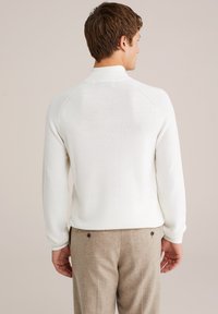 Weißer Ripp-Pullover mit hohem Kragen, getragen mit beigen Hosen, die über eine dezente Textur und Knopfdetails an der Taille verfügen.