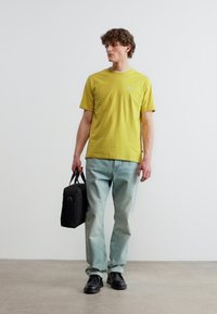 Άνδρας που φοράει κίτρινη μπλούζα, ανοιχτό μπλε τζιν, μαύρα παπούτσια, κρατώντας μαύρη τσάντα, στέκεται σε γκρι δάπεδο μπροστά σε απλό φωτεινό τοίχο.