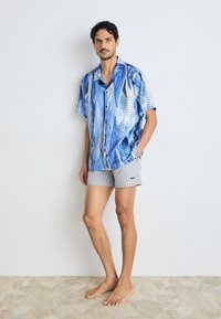 Short-sleeve button-up shirt in blue with abstract patterns, paired with gray striped shorts featuring a logo. Barefoot on patterned floor.