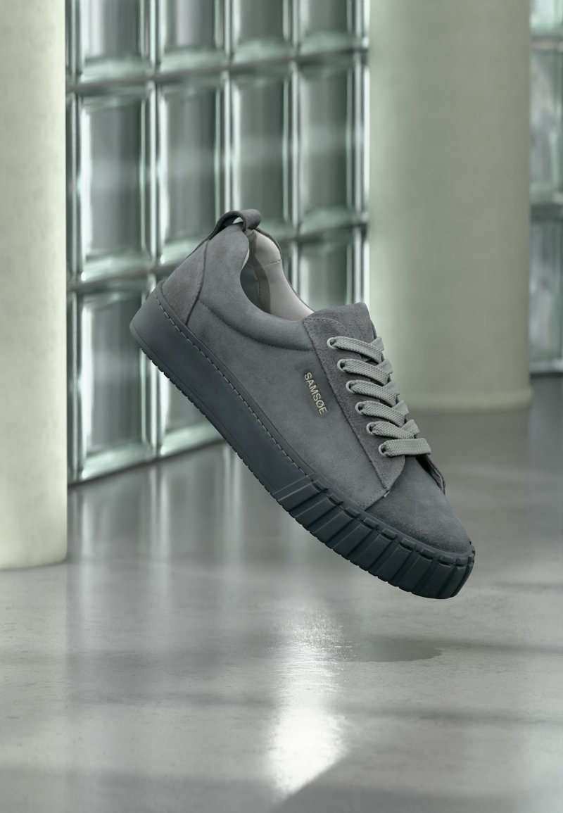 Γκρι sneaker σουέτ με μαύρη καουτσούκ σόλα και κορδόνια, με την επωνυμία "SAMSOE", που αιωρείται σε εσωτερικό χώρο κοντά σε τοίχο από γυάλινα τουβλάκια και δάπεδο από σκυρόδεμα.