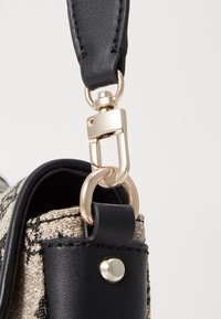 Cinghia in pelle nera attaccata con una clip metallica dorata a una borsa in tessuto beige strutturato con un motivo nero. Finitura liscia, dettagli hardware robusti.