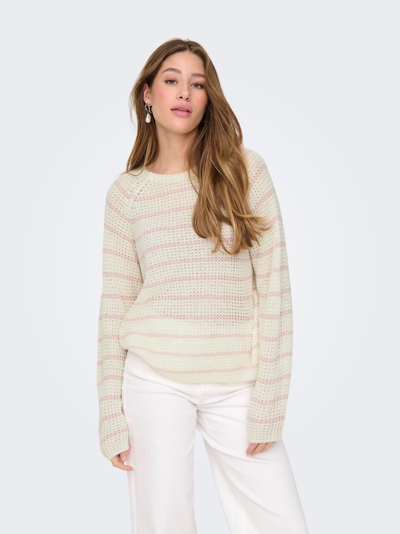 Femme aux longs cheveux bruns portant un pull en maille rayé crème et rose et un pantalon blanc, debout devant un fond clair uni.