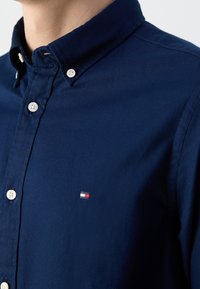 Chemise bleu marine à boutons avec col pointu, boutons blancs et un petit logo rouge, blanc et bleu sur la poitrine. Tissu en coton avec une finition texturée.