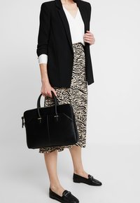Kvinde iført sort blazer, hvid bluse, midi-nederdel i dyreprint, sorte loafers, der holder en sort læder arbejdstaske ved siden af.