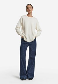 Cremiger Pullover mit weiten Ärmeln, gerippten Bündchen und Saum. Kombiniert mit dunkelblauen weiten Jeans und schwarzen Absatzschuhen.
