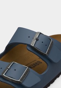 Κοντινή λήψη των μπλε λουριών σανδαλιού Birkenstock με μεταλλικά αγκράφες και ορατή καφέ σόλα κάτω από αυτά.