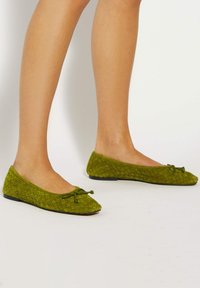Green woven flats made of soft material, featuring a rounded toe and a decorative bow at the front. Low-profile black sole.