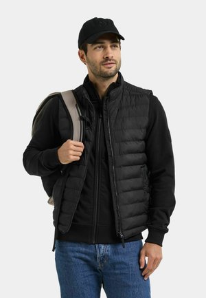 Man met zwarte pet, zwarte gewatteerde bodywarmer en jeans staat, houdt een rugzakriem vast en kijkt opzij met een neutrale uitdrukking.