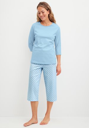 Donna in piedi a piedi nudi, indossa una maglia a maniche lunghe azzurro chiaro e pantaloni corti azzurro chiaro con motivo a pois bianchi.