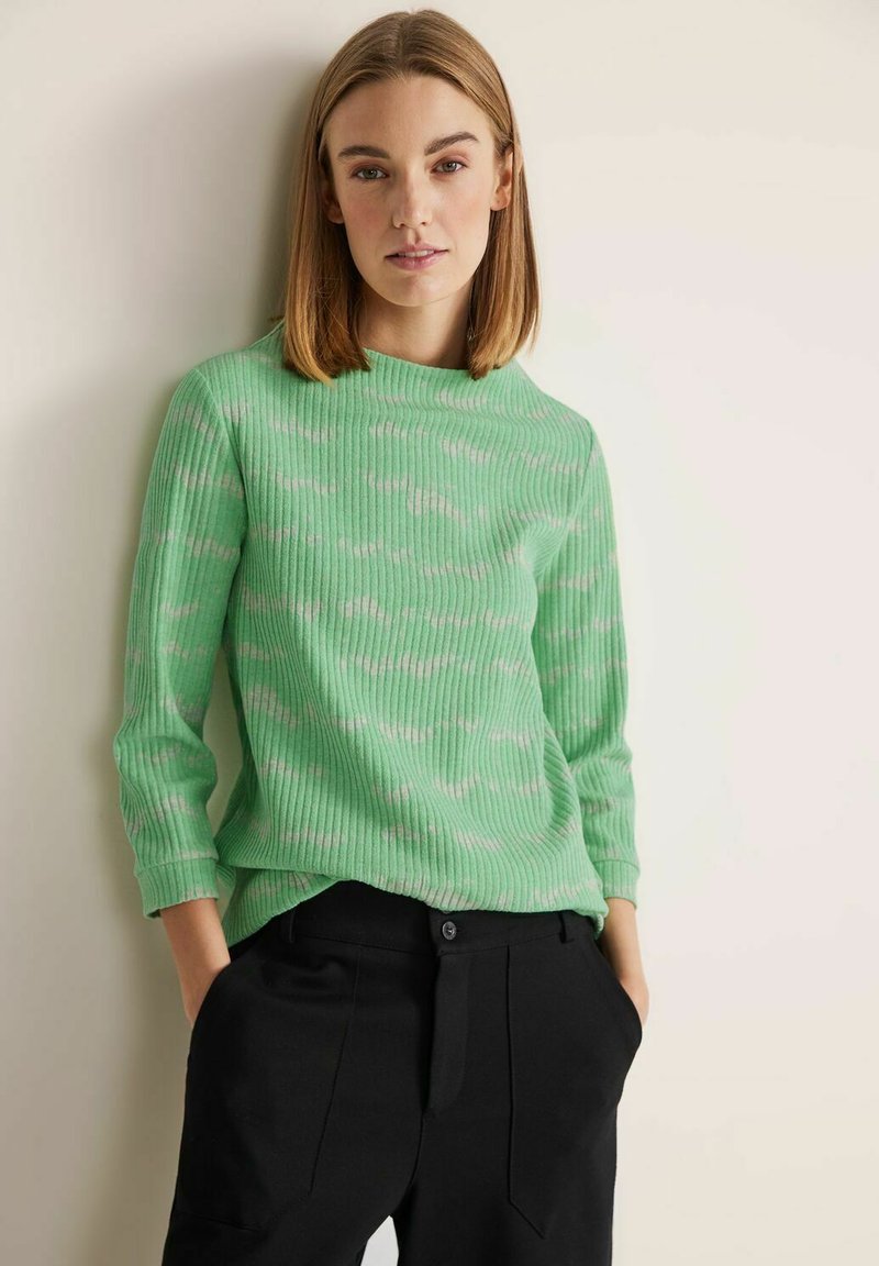 Maglione verde a coste con un collo alto e un sottile motivo crema simile a nuvole, indossato con pantaloni neri a gamba affusolata.