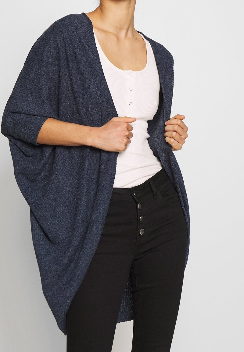 Frau trägt eine dunkelblaue strukturierte Strickjacke über einem weißen, knöpfbaren Tanktop und hochtaillierten schwarzen Hosen mit vier Knöpfen auf der Vorderseite.