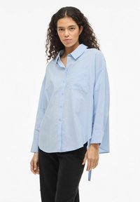 Chemise bleu clair en coton, à manches longues, avec une poche poitrine et une coupe décontractée agrémentée d'un lien sur le côté.