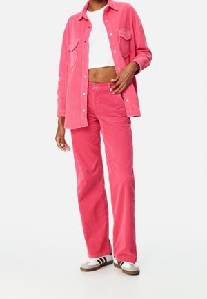 Personne portant un crop top blanc, une chemise et un pantalon en velours côtelé rose, ainsi que des baskets blanches avec des rayures noires, debout devant un fond uni.