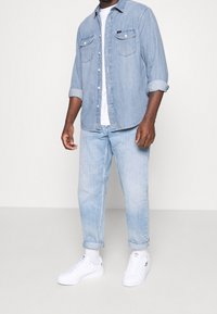 Světlá modrá džínová košile, bílé tričko a světlé modré džíny, doplněné bílými teniskami. Košile má dvě kapsy na prsou a podhrnuté rukávy.