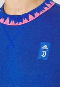 Gros plan d’un t-shirt Adidas bleu avec un motif triangulaire rose sur le col et un écusson du logo Juventus sur la poitrine.