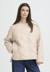 Veste matelassée beige avec un col rond, présentant un motif texturé et une coupe décontractée. Le modèle la porte avec un jean bleu foncé.