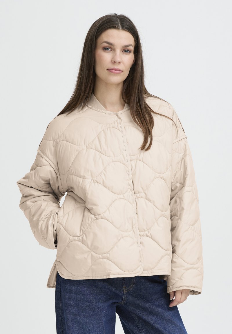 Veste matelassée beige avec un col rond, présentant un motif texturé et une coupe décontractée. Le modèle la porte avec un jean bleu foncé.