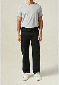 Homme portant un t-shirt gris clair à manches courtes, un pantalon noir à jambes droites et des baskets blanches, debout devant un fond clair uni.